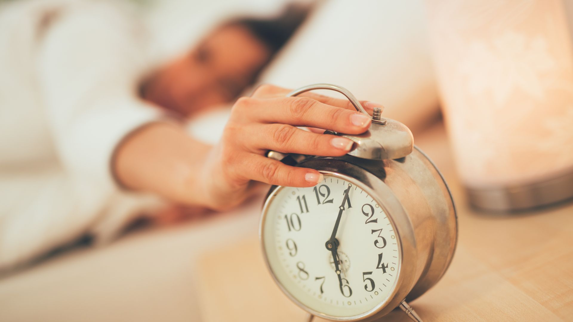 A woman reaches from her bed to turn off her alarm clock.